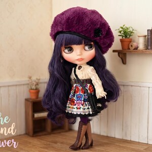 May include: A doll with long wavy purple hair wearing a dark purple fuzzy beret, a cream-colored lace long-sleeve top, and a skirt featuring a colorful floral pattern. The doll has a black crossbody bag and brown lace-up boots.