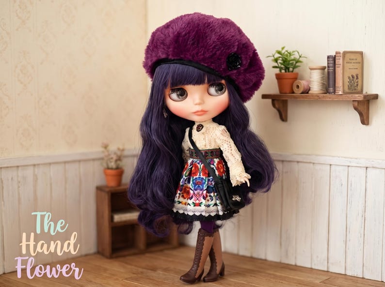 May include: A doll with long wavy purple hair wearing a dark purple fuzzy beret, a cream-colored lace long-sleeve top, and a skirt featuring a colorful floral pattern. The doll has a black crossbody bag and brown lace-up boots.