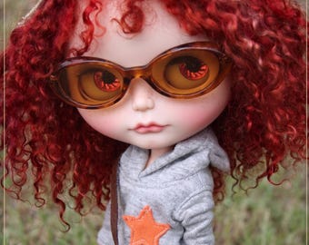 May include: A doll with red curly hair, wearing brown-framed sunglasses, a gray hoodie with an orange star, and a camouflage cap. The doll has a small camouflage purse with a brown strap. The doll is standing in a grassy area.
