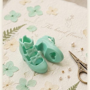 Op de afbeelding: Een paar mintgroene miniatuur high-heels met strikaccenten. De schoenen staan op een getextureerd oppervlak met gedroogde bloemen, parels en kleine gouden kralen. De tekst "The hand flower" is zichtbaar.