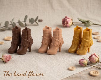 Puede incluir: Tres pares de botines en miniatura en marrón oscuro, beige claro y amarillo mostaza. Cada par presenta un detalle de flecos alrededor del tobillo, punta afilada y tacón alto. El texto "The hand flower" aparece en la esquina inferior izquierda.
