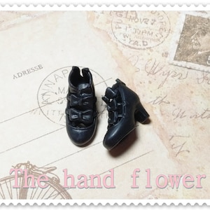 Op de afbeelding: Twee miniatuur zwarte pumps met strikaccenten. De schoenen worden getoond op een vintage-achtige achtergrond met postzegels en handschrift. De tekst "The hand flower" is zichtbaar aan de onderkant.