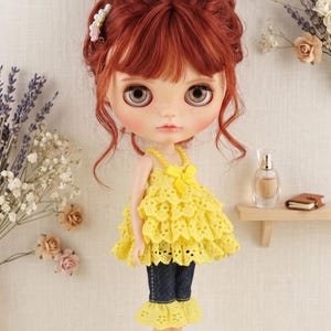 May include: A doll with auburn hair styled in an updo, wearing a yellow eyelet lace tiered sleeveless top with a bow, dark blue denim capri trousers with yellow eyelet lace ruffles at the hem, and yellow shoes. The text The hand flower is visible.