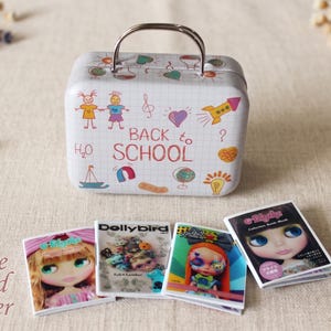 Puede incluir: Una fiambrera de metal blanco con asa plateada, decorada con ilustraciones coloridas con temática escolar y las palabras "BACK TO SCHOOL". Cuatro revistas de muñecas en miniatura están delante de la fiambrera.