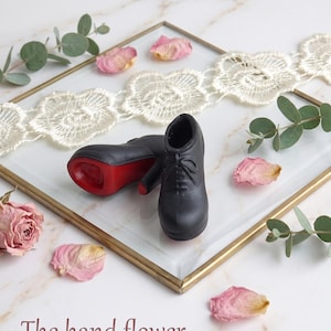 May include: A pair of miniature black lace-up shoes with red soles. The shoes are displayed on a glass tray with a gold frame, decorated with white lace trim, dried rose petals, and green eucalyptus sprigs. The text "The hand flower" is visible.