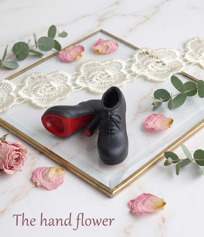 May include: A pair of miniature black lace-up shoes with red soles. The shoes are displayed on a glass tray with a gold frame, decorated with white lace trim, dried rose petals, and green eucalyptus sprigs. The text "The hand flower" is visible.