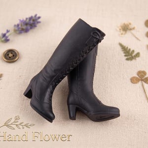 May include: A pair of black knee-high boots with a small heel, featuring decorative stitching and a lace-up design. The boots are displayed on a neutral surface with dried flowers and the text "The Hand Flower".