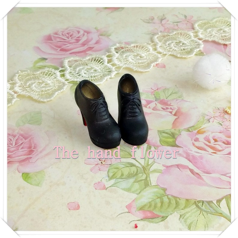 May include: A pair of miniature black lace-up high-heeled shoes with a rounded toe and a red sole detail. The shoes feature molded laces on the front and a smooth finish. The text "The hand flower" is printed in pink letters below the shoes.