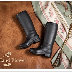 May include: A pair of black leather riding boots with laces, presented on a brown leather surface. A riding crop and a floral scarf are also visible. The Hand Flower logo is present.