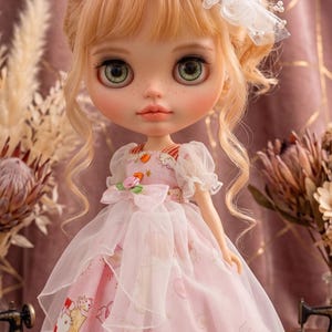 May include: A doll with green eyes and blonde hair, wearing a pink dress with a Hello Kitty print. The dress has a white overlay and a bow at the waist. The doll has a white headpiece with feathers and pearls. The text "Thehand flower" is at the top.