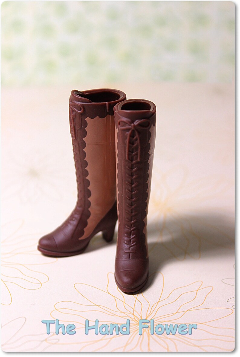 May include: A pair of brown knee-high boots featuring a two-tone design with a darker brown toe, heel, and lace-up front panel, contrasted by a lighter brown side panel with a scalloped edge. The text "The Hand Flower" is printed below.