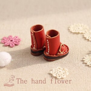 May include: Miniature red leather boots with white stitching and gold buckles. The boots are paired with white lace doilies, a pink crocheted flower, and a white pom-pom. The text "The hand flower" is at the bottom.
