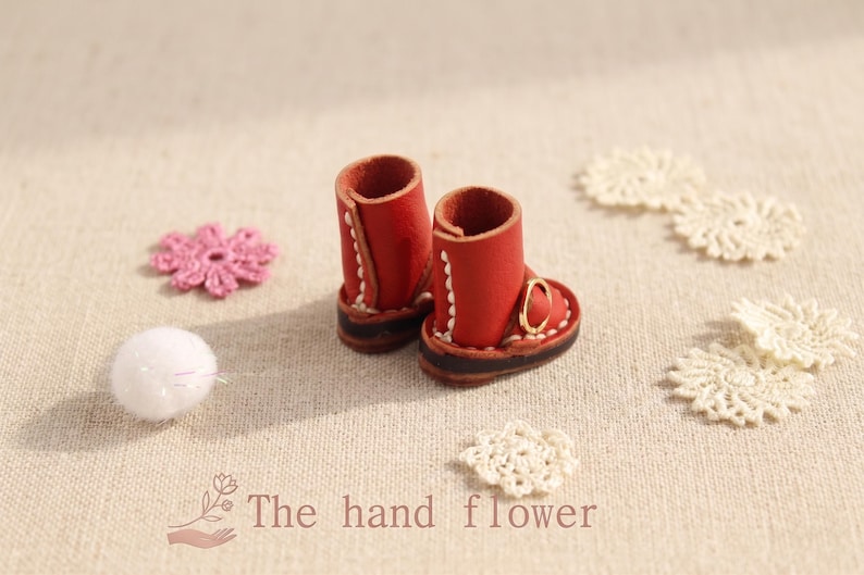 May include: Miniature red leather boots with white stitching and gold buckles. The boots are paired with white lace doilies, a pink crocheted flower, and a white pom-pom. The text "The hand flower" is at the bottom.