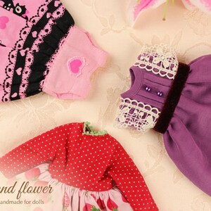 May include: A collection of doll clothes in various styles and colors. Includes a red polka-dot top, a pink dress with strawberry print, and a purple dress. Other items include a pink scarf with black cat and heart patterns, and a purple top with lace trim.