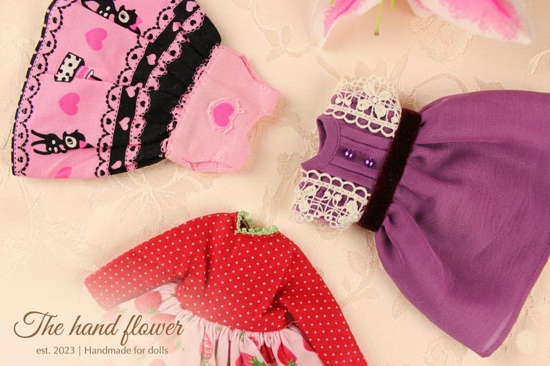 May include: A collection of doll clothes in various styles and colors. Includes a red polka-dot top, a pink dress with strawberry print, and a purple dress. Other items include a pink scarf with black cat and heart patterns, and a purple top with lace trim.