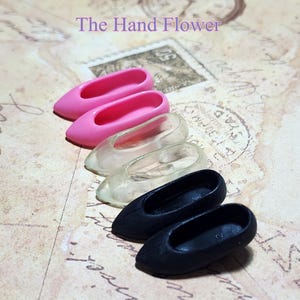 Puede incluir: Una colección de zapatos de muñeca en miniatura en rosa, transparente y negro. Los zapatos están dispuestos en fila, con el texto "The Hand Flower" en morado encima. El fondo es un papel de estilo vintage.
