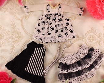 May include: Three doll outfits are displayed on a floral patterned surface. One outfit is a white dress with black paw prints and a pearl necklace. Another is a black skirt with white stripes. The third is a layered skirt with polka dots and lace. The text "The hand flower" is visible.