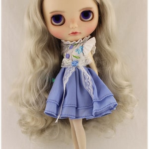 May include: A doll with long, silver hair wearing a blue dress with white lace trim and a floral print top. The doll is wearing white tights and white shoes.