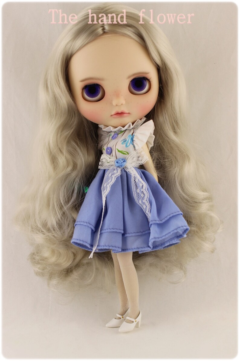 May include: A doll with long, silver hair wearing a blue dress with white lace trim and a floral print top. The doll is wearing white tights and white shoes.