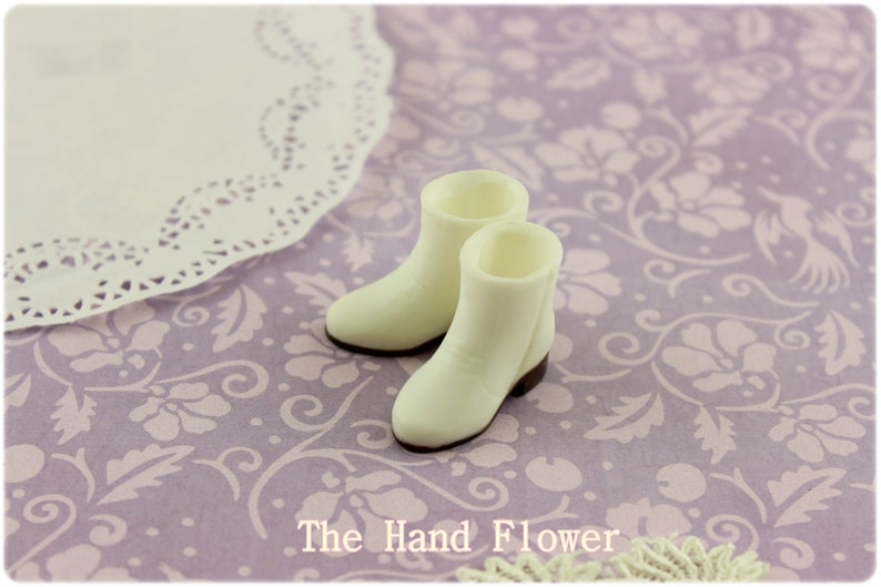 Puede incluir: Un par de botas de vaquero blancas en miniatura con suelas y tacones marrones. Est&aacute;n sobre una superficie con estampado floral lavanda, con un tapete de encaje blanco. El texto "The Hand Flower" es visible.