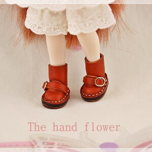 May include: Close-up of doll's legs wearing red leather boots with gold buckles. The boots have a stitched sole and a strap detail. The text "The hand flower" is at the bottom of the image.