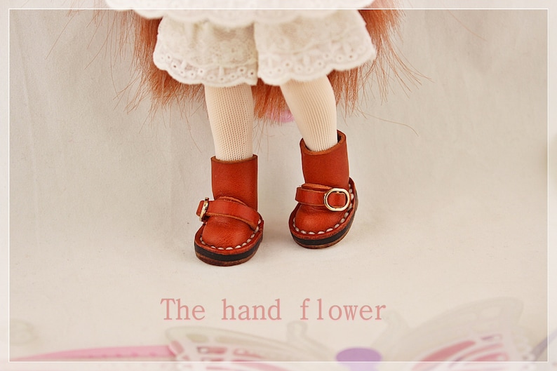 May include: Close-up of doll's legs wearing red leather boots with gold buckles. The boots have a stitched sole and a strap detail. The text "The hand flower" is at the bottom of the image.