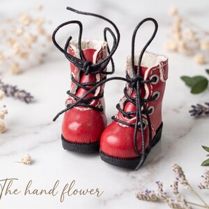 May include: A pair of miniature red boots with black laces. The boots have white trim and a black sole. The image includes dried flowers and the text "The hand flower".