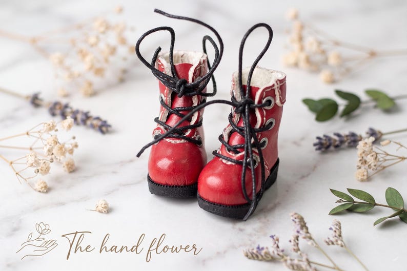 May include: A pair of miniature red boots with black laces. The boots have white trim and a black sole. The image includes dried flowers and the text "The hand flower".