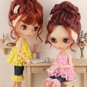 May include: Two dolls with auburn hair styled in updos, one wearing a yellow ruffled top and dark denim capris, the other in a pink ruffled top and denim capris, sitting on a wooden stool. The text "The hand flower" is printed at the bottom.