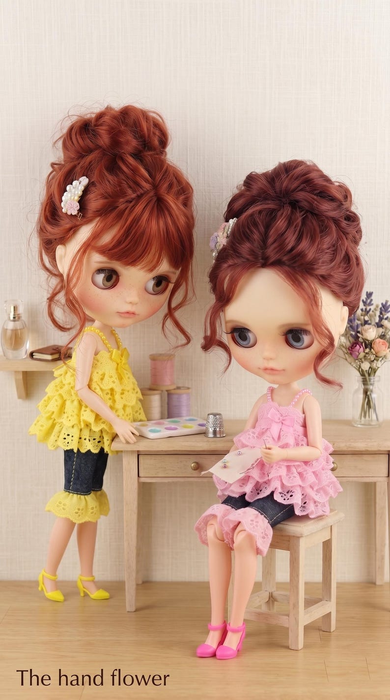 May include: Two dolls with auburn hair styled in updos, one wearing a yellow ruffled top and dark denim capris, the other in a pink ruffled top and denim capris, sitting on a wooden stool. The text "The hand flower" is printed at the bottom.
