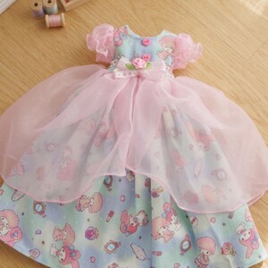 May include: A doll dress featuring a light blue and pink color scheme. The dress has a layered design with a printed pattern of cartoon characters. It includes a bow, pink buttons, and a sheer overlay. The dress is approximately 10 inches in length.