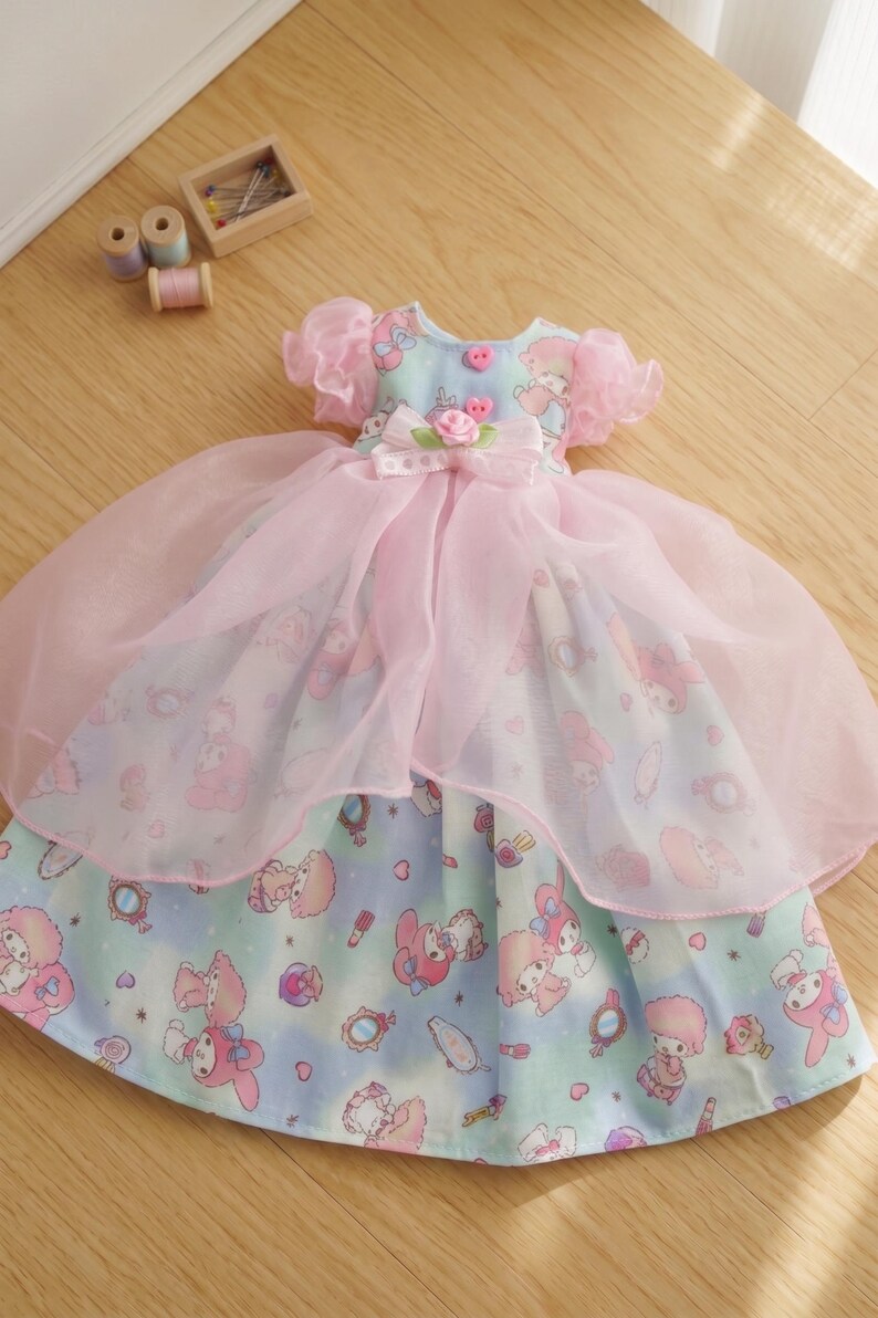 May include: A doll dress featuring a light blue and pink color scheme. The dress has a layered design with a printed pattern of cartoon characters. It includes a bow, pink buttons, and a sheer overlay. The dress is approximately 10 inches in length.