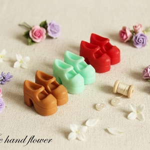 Puede incluir: Tres pares de zapatos de plataforma en miniatura en marrón, verde menta y rojo, acompañados de pequeñas flores artificiales rosas, moradas y blancas, un carrete de madera con hilo blanco y dos botones blancos, con el texto The hand flower.