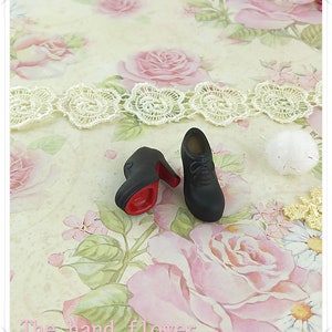 May include: A pair of miniature black lace-up shoes with high heels and a bright red sole. The shoes feature a rounded toe and molded laces on the upper surface. The text "The hand flower" is printed in light pink at the bottom of the image.