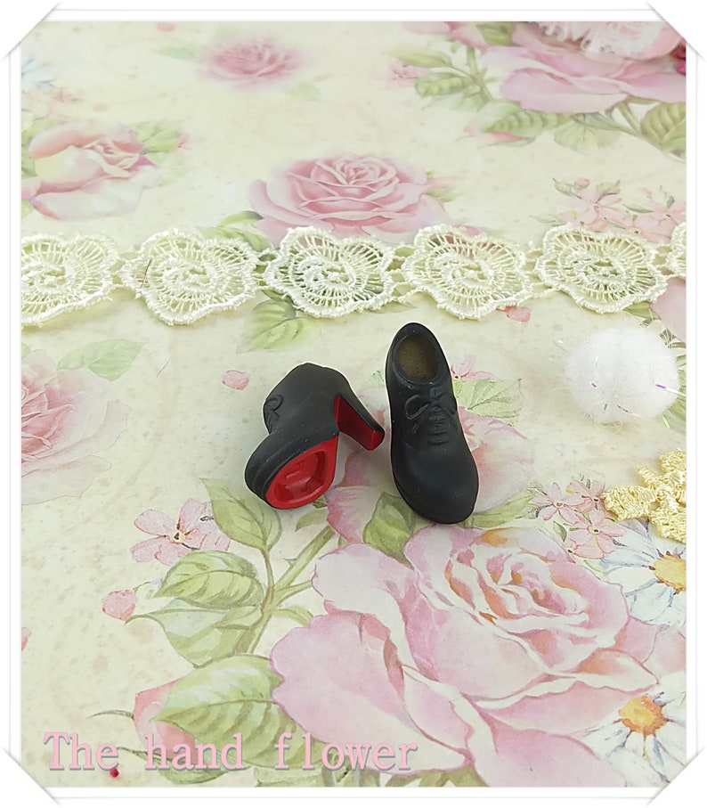 May include: A pair of miniature black lace-up shoes with high heels and a bright red sole. The shoes feature a rounded toe and molded laces on the upper surface. The text "The hand flower" is printed in light pink at the bottom of the image.