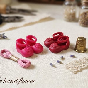 May include: A pair of bright pink doll shoes and a pair of dark red doll shoes, each featuring a molded flower detail on the toe and an ankle strap. Also included are a small brass thimble and a coiled length of pink embroidery thread.