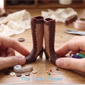 May include: A pair of miniature doll boots in light brown and dark brown, featuring a lace-up front design with scalloped edges, a small heel, and a tall shaft. The boots are displayed alongside sewing notions including buttons and pins.