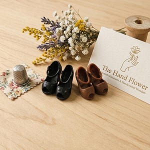 Könnte beinhalten: Auf einer Holzoberfläche sind zwei Paar Miniatur-Mary-Jane-Schuhe, schwarz und braun, angeordnet. Ein kleiner Strauß Trockenblumen, ein Fingerhut und eine Karte mit dem Text "The Hand Flower" sind ebenfalls vorhanden. Ideal für ein Puppenhaus.