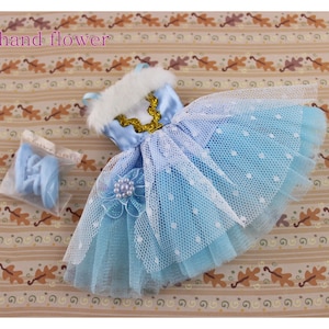 May include: A light blue tulle dress with white polka dots and a white fur trim around the neckline. The dress has a gold trim and a white flower with a pearl accent. The dress is on a brown background with a pattern of leaves.