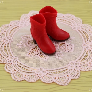May include: A pair of miniature red boots with a textured design, resting on a pink lace doily. The boots have small heels and are set against a light yellow background.