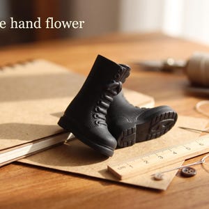 May include: A pair of miniature black lace-up boots with textured soles, positioned next to a wooden ruler showing measurements in centimeters. The text "The hand flower" is visible in the upper left corner of the image.