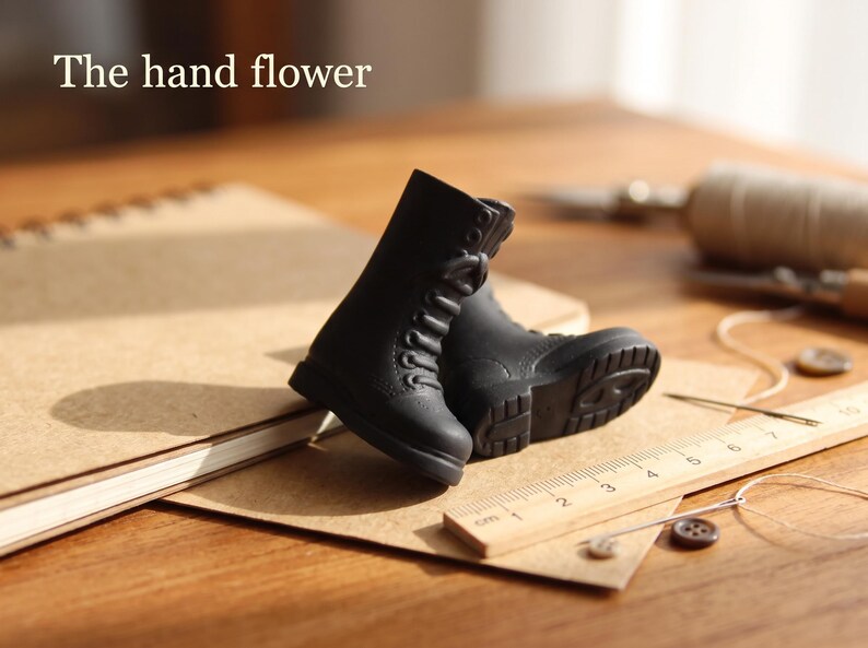 May include: A pair of miniature black lace-up boots with textured soles, positioned next to a wooden ruler showing measurements in centimeters. The text "The hand flower" is visible in the upper left corner of the image.
