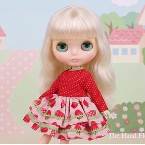 May include: A doll with long, blonde hair and large, blue eyes. The doll wears a red, long-sleeved top with white polka dots and a pink tiered skirt with strawberry patterns. The background features a painted landscape.