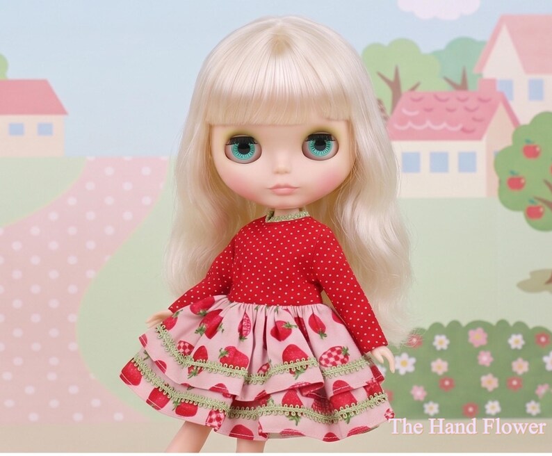 May include: A doll with long, blonde hair and large, blue eyes. The doll wears a red, long-sleeved top with white polka dots and a pink tiered skirt with strawberry patterns. The background features a painted landscape.