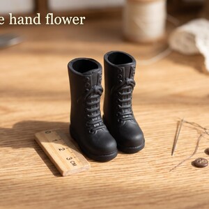May include: A pair of miniature black lace-up boots with a matte finish. The boots are positioned next to a small wooden ruler showing a scale of 3 cm. The text "The hand flower" is visible in the upper left corner of the image.