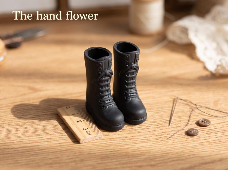 May include: A pair of miniature black lace-up boots with a matte finish. The boots are positioned next to a small wooden ruler showing a scale of 3 cm. The text "The hand flower" is visible in the upper left corner of the image.