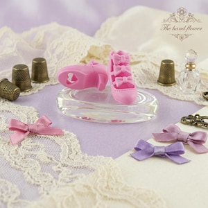 Op de afbeelding: Een paar miniatuur roze pumps met strikaccenten, gepresenteerd op een helder glazen ovaal. De schoenen staan naast een vingerhoed, een kleine parfumfles en decoratieve strikken. De achtergrond heeft kant en een bloemenpatroon.