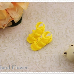 May include: A pair of bright yellow miniature sandals. The sandals have a strappy design with a rounded toe and a small heel. The sandals are made of a smooth, solid material. The background includes a floral arrangement and a small bear figurine.