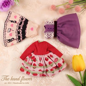 May include: Doll dresses in various styles. One dress is red with white polka dots and a strawberry-patterned skirt. Another is a light pink dress with black cat and heart details. A purple dress with lace trim is also shown.