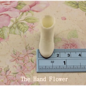 Puede incluir: Una peque&ntilde;a bota de color crema, de unos 3,2 cm de altura, se muestra sobre una superficie con estampado floral. La bota tiene una punta redondeada y un peque&ntilde;o tac&oacute;n. El fondo presenta un dise&ntilde;o floral vintage con rosas rosas y hojas verdes.